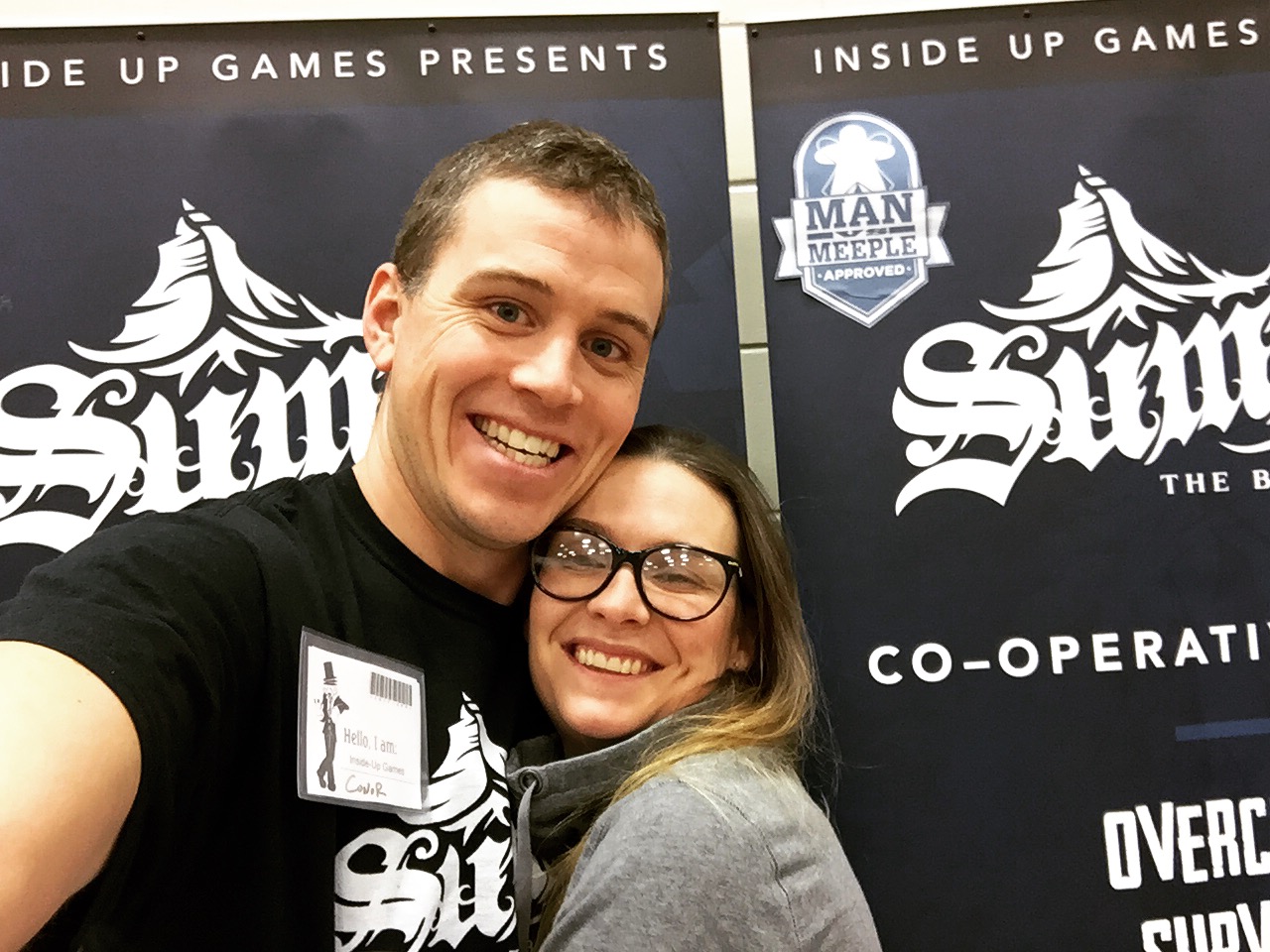 Conor McGoey hugging a femme presenting person in front of a banner for his popular Summit Board Game