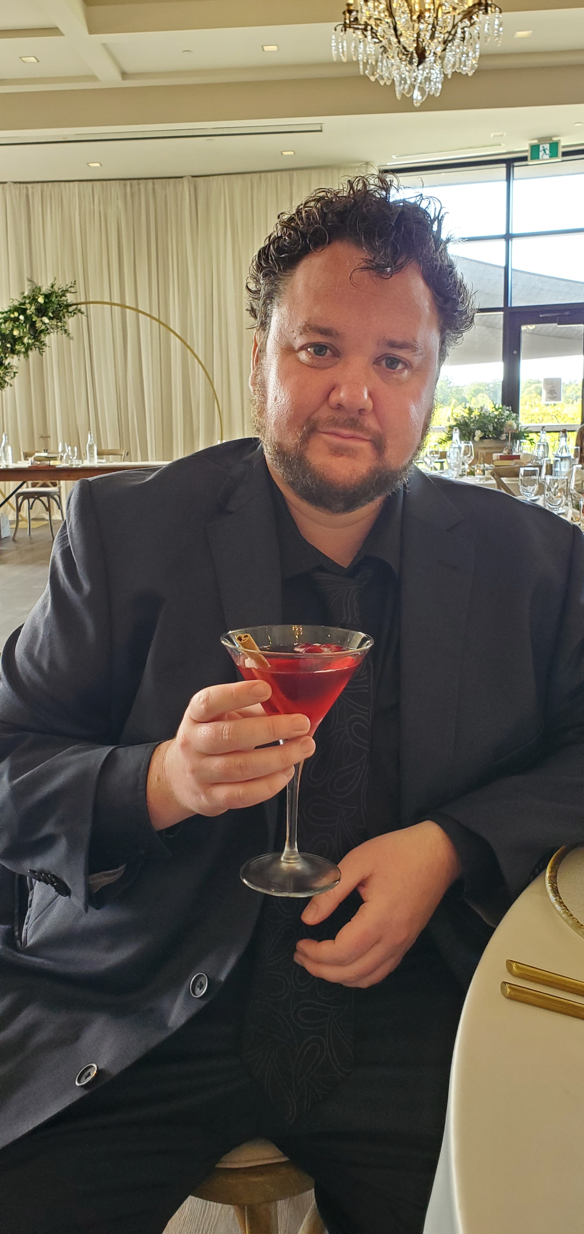 Daniel Legault, in a black suit and lie, leaning against a table, while saluting the camera with cocktail in a martini glass with a cinnamon stick garnish.