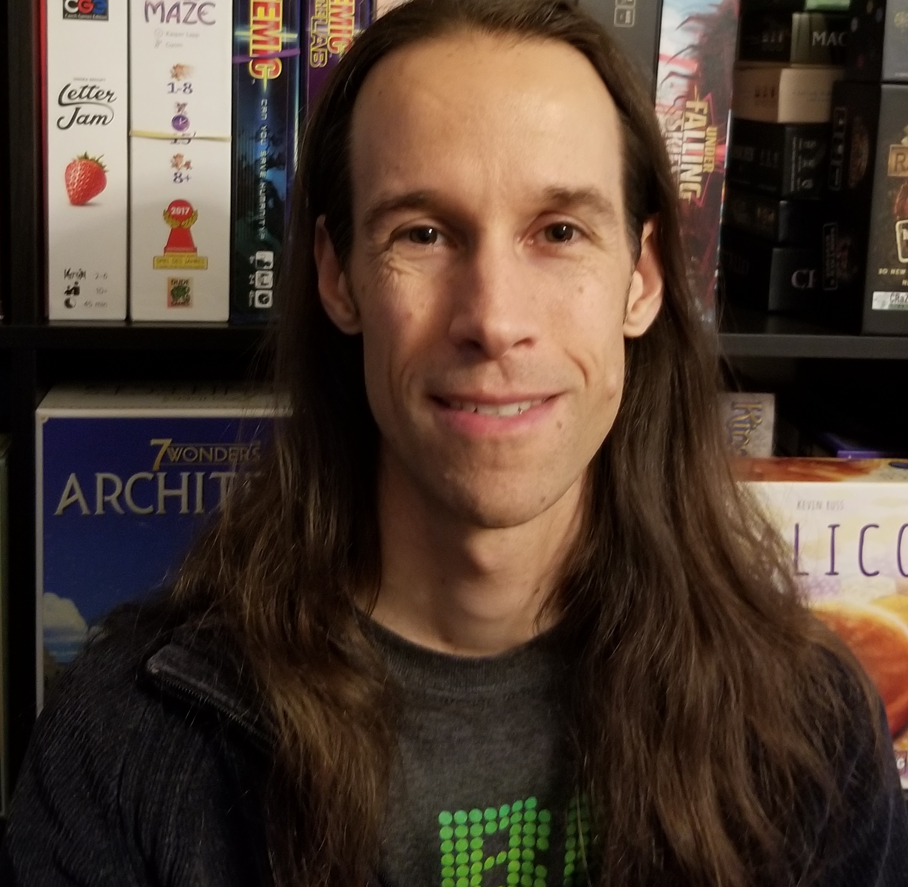 Joe Slack smiling into the camera in front of a shelf full of boardgames like pandemic, 7 Wonders and more