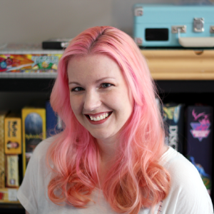 Shannon, vibrant pink hair catching the light, smiles in front of a shelf of games