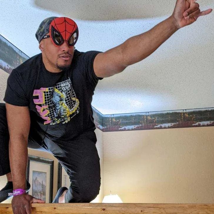 Ricky, wearing a paper half-mask of Spider-man's cowl, kneels precariously on a bannister, holding his hand out with his pinky and index finger extended, and his middle and ring fingers tucked into his hand, using an imaginary web-shooter.