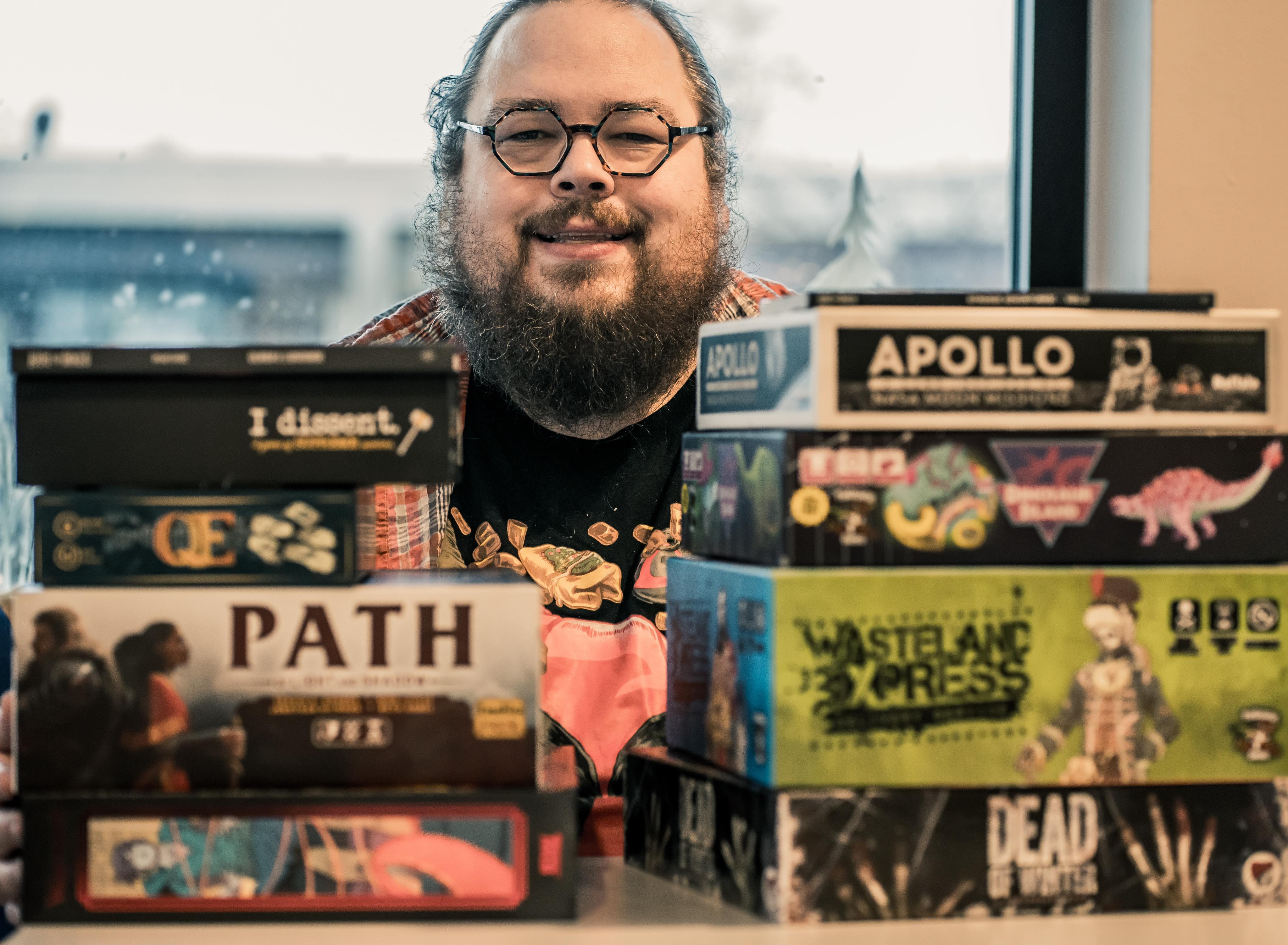 Jon sitting behind a stack of games he's designed including Dead of Winter, Dinosaur Island, Wasteland Express and More