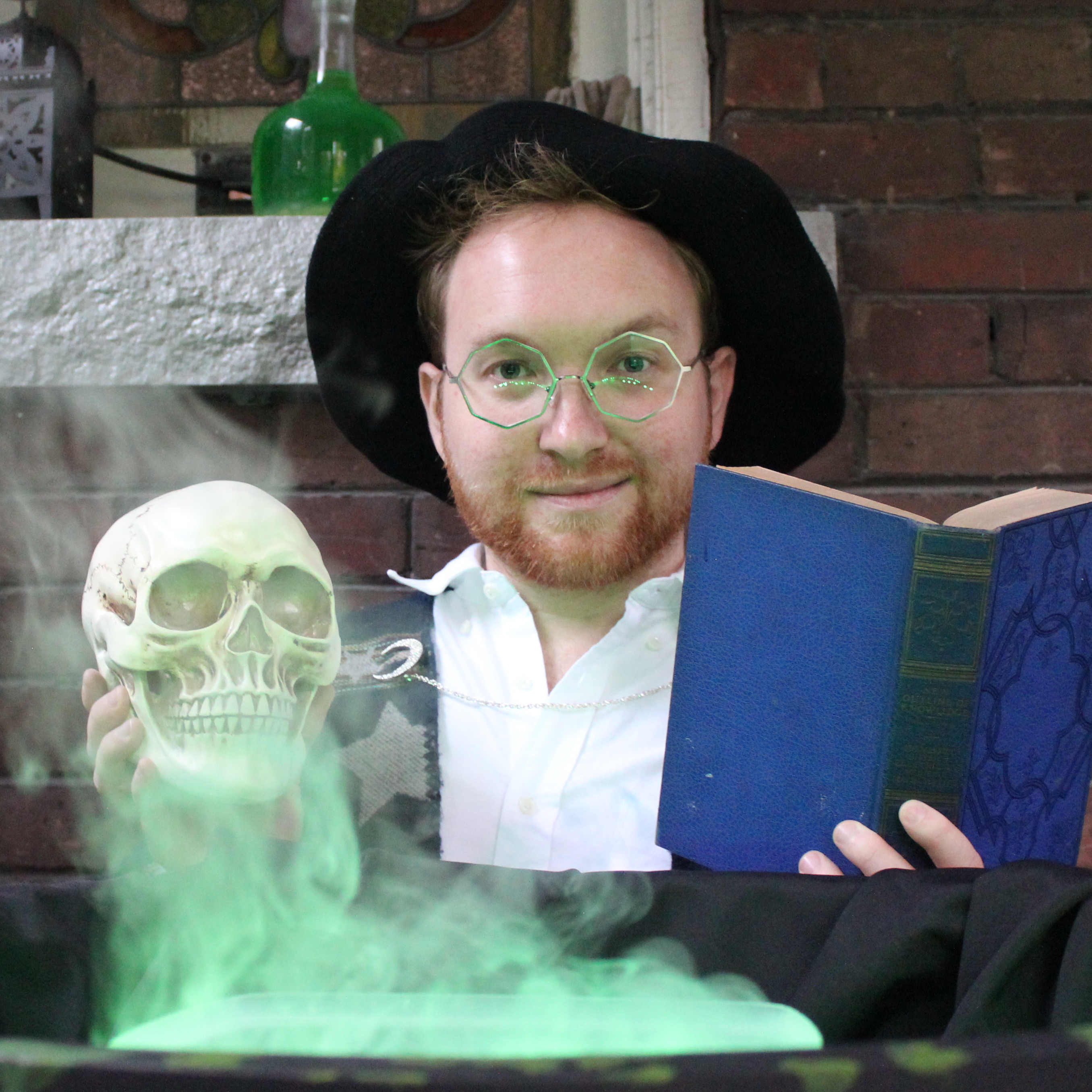 Jesse, sits in front of a cauldron that belches smoke and flows green, he holds a skull in his right hand and a tome in his left, and smiles impishly in a black velvet hat.