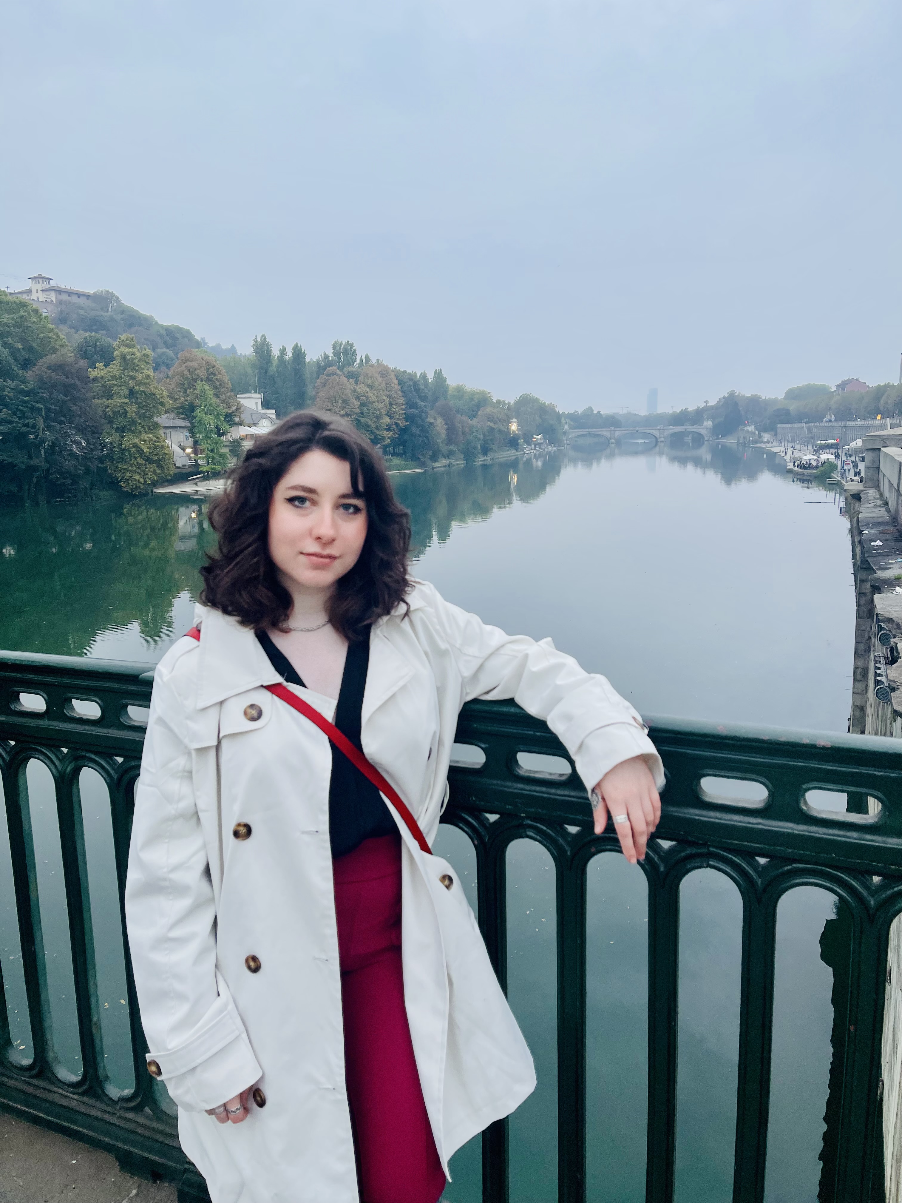 Tatiana in a white overcoat leans casually against a metal fence on a bridge overlooking a picturesque canal