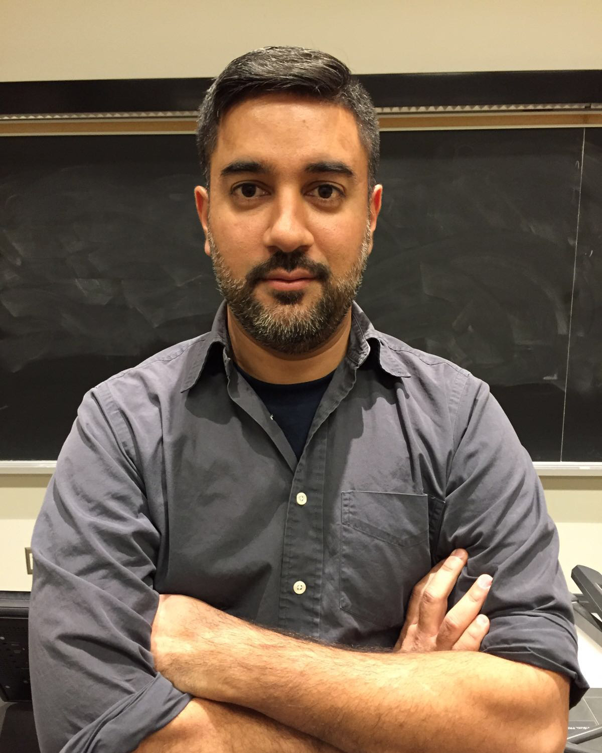Shiraz wearing a blue collared shirt, with arms folded in front of him, standing in front of a recently cleaned blackboard