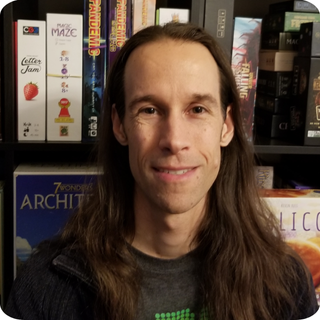 Photo of Joe Slack standing in front of a bookshelf of board games.