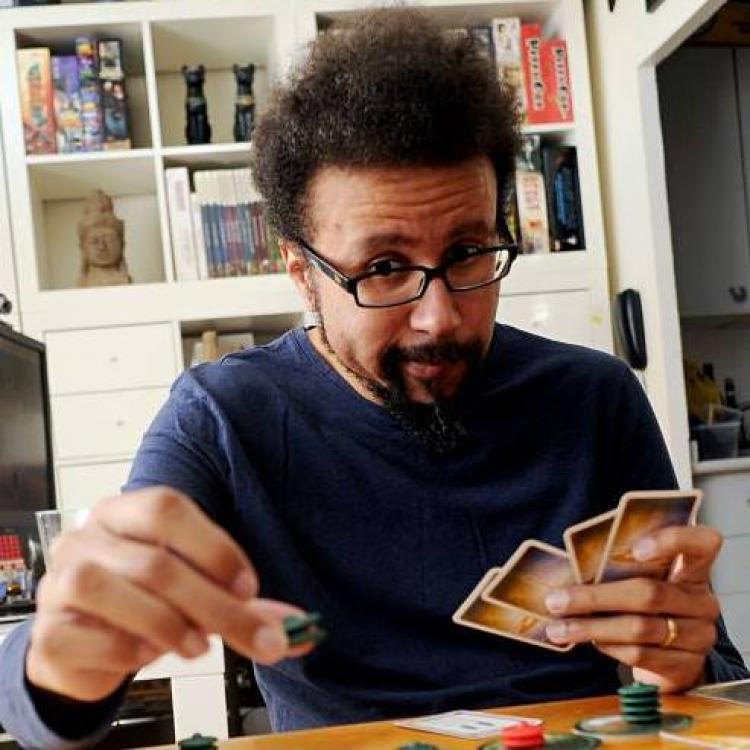 Eric Lang holds game cards in his left hang, while placing two green discs in his hand - it looks like he's playing cosmic encounters, but his facial expression suggests he might not be winning