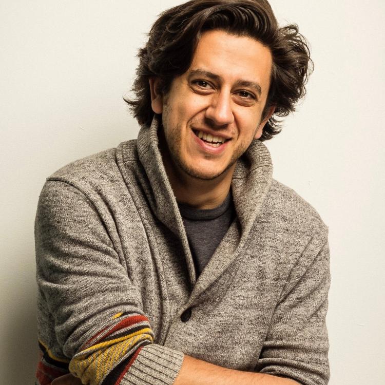 Kris Siddiqi wearing a finely knit sweater, smiling effortlessly at the camera