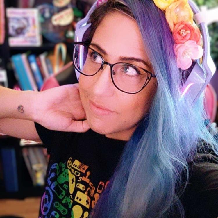 Person wearing glasses, wearing a shirt that has multiple video-game type controllers on it, wearing a rainbow flower crown