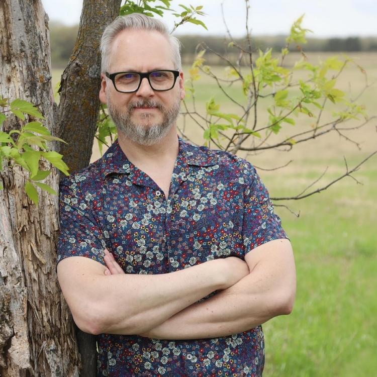 Greg Leatherman in sleek black glasses, leaning casually against a tree