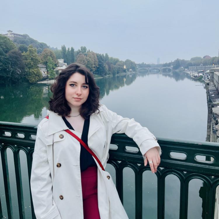 Tatiana in a white overcoat leans casually against a metal fence on a bridge overlooking a picturesque canal