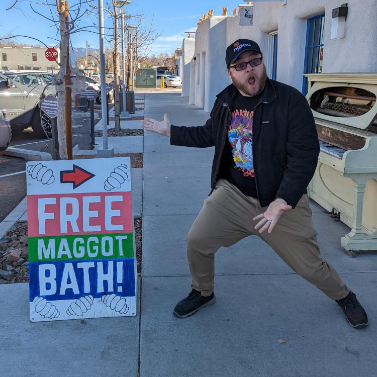 Graham standing on the sidewalk, gesturing fervently to the camera and a sandwich board all at once. The sandwich board says "Free Maggot Bath" and the expression on Graham's face is a unique combination of horror, surprise, exhultation, and curiosity. 