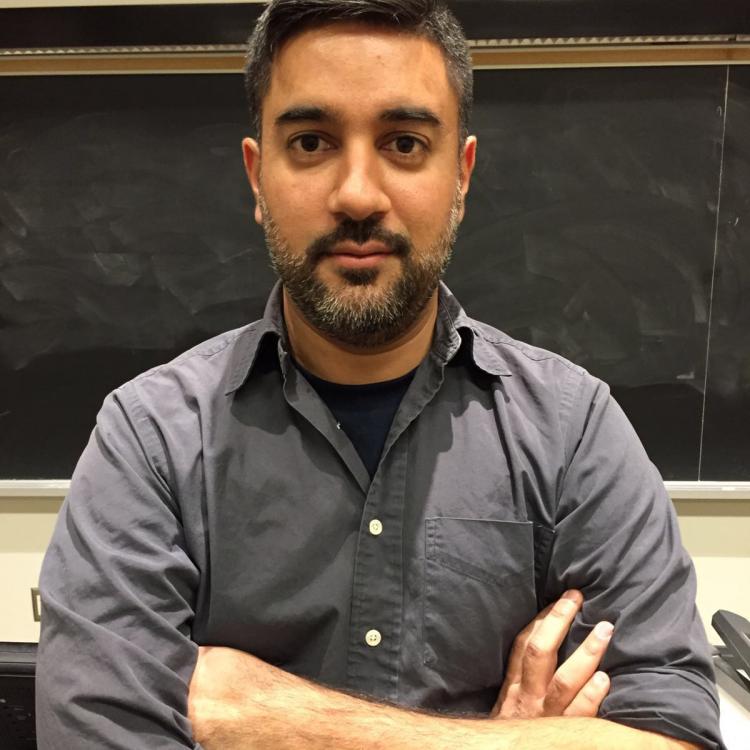 Shiraz wearing a blue collared shirt, with arms folded in front of him, standing in front of a recently cleaned blackboard